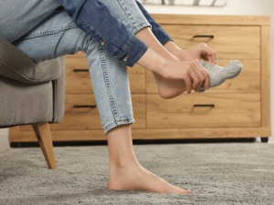 Person sits barefoot on a rug, putting on a gray sock with a wooden dresser in the background.